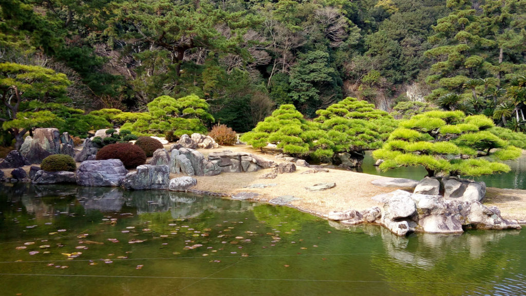 Clipped pines and weathered rocks on a sandy shore of a still green pond in a Japanese garden, with a wooded hillside rising behind.