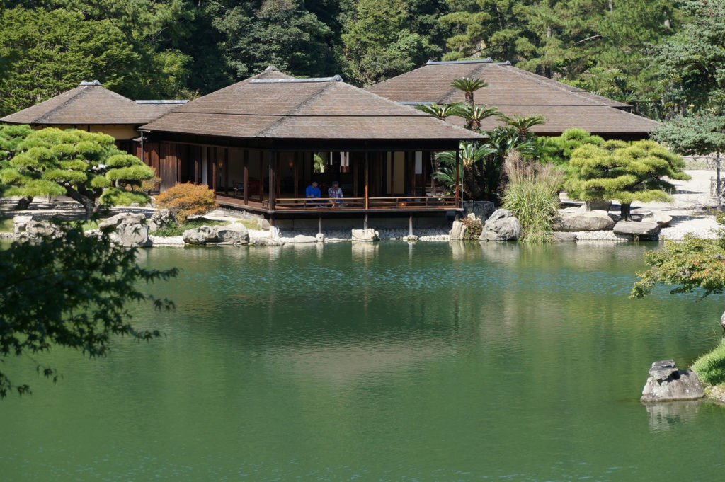 A traditional teahouse with a deep tiled roof sits at the water’s edge of a green pond, with visitors resting on its veranda among sculpted pines.