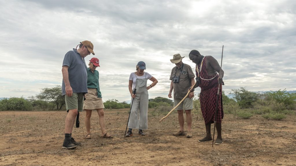 Guests on a walking safari in Shompole Conservancy watch a guide point out tracks in the dry earth under an overcast sky.