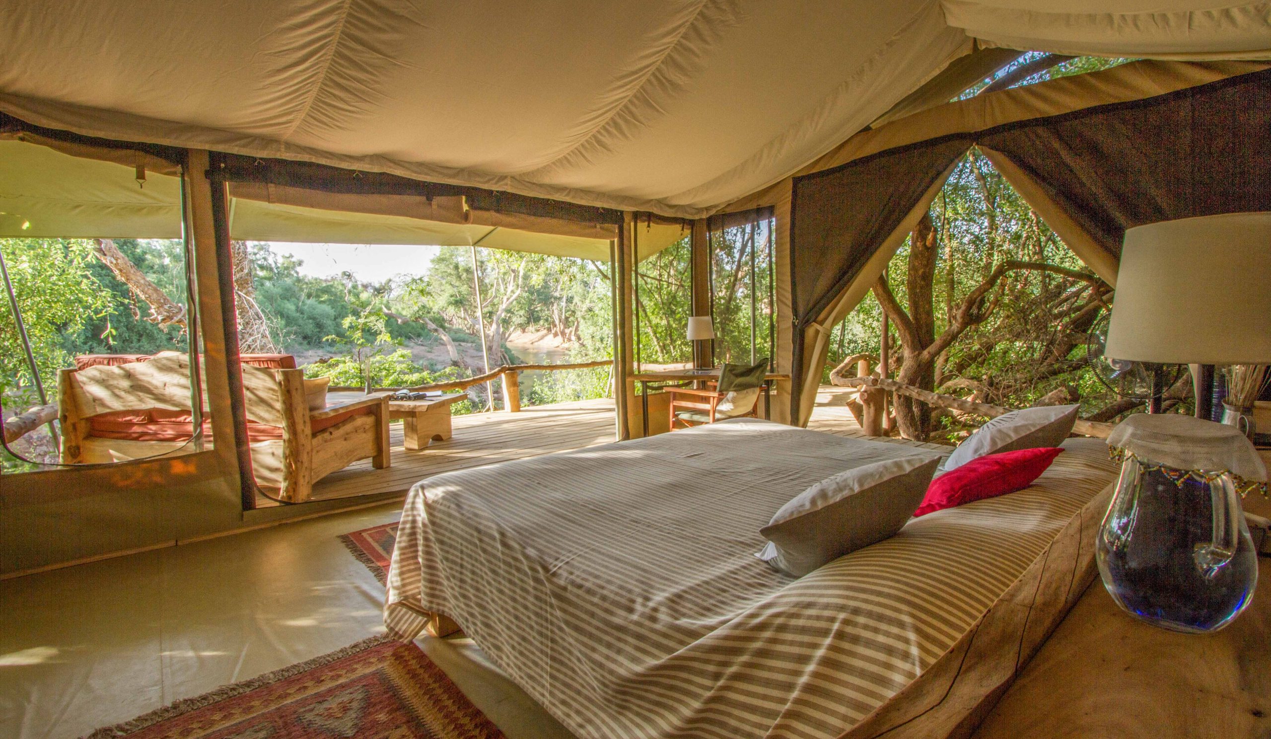 Double tent at Shompole Wilderness opening onto a timber deck with a sofa and armchair overlooking the river in warm light.