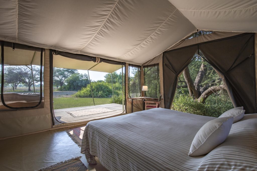 Double bed with striped linen and a writing desk inside a canvas tent, its mesh doors rolled up onto a deck and acacia woodland.