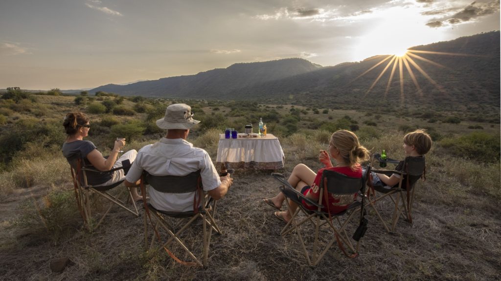 Guests and two children in camp chairs at a sundowner, with drinks on a table and the sun setting behind a ridge in southern Kenya.