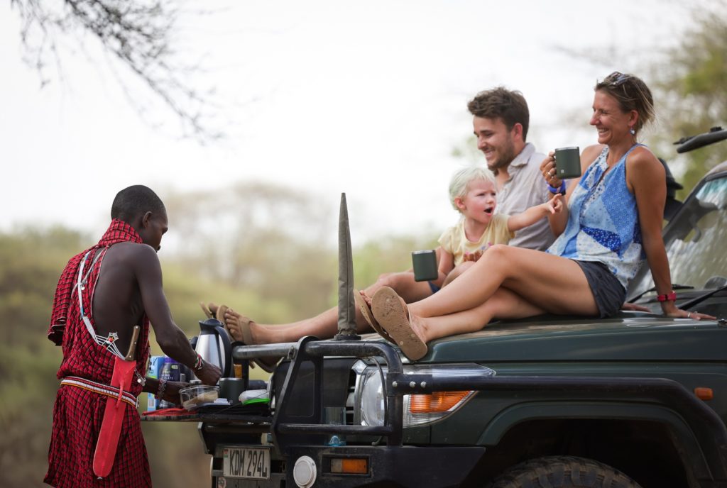 Guide pours hot drinks from the bull bar of a four-wheel drive while guests and a small child sit on the bonnet on a game drive.