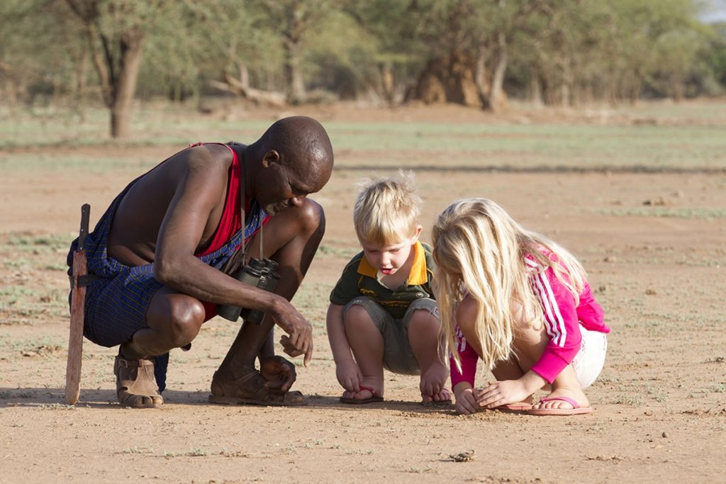 Guide crouches beside two children on a tracking lesson, studying animal prints in the sandy soil of Shompole Conservancy.