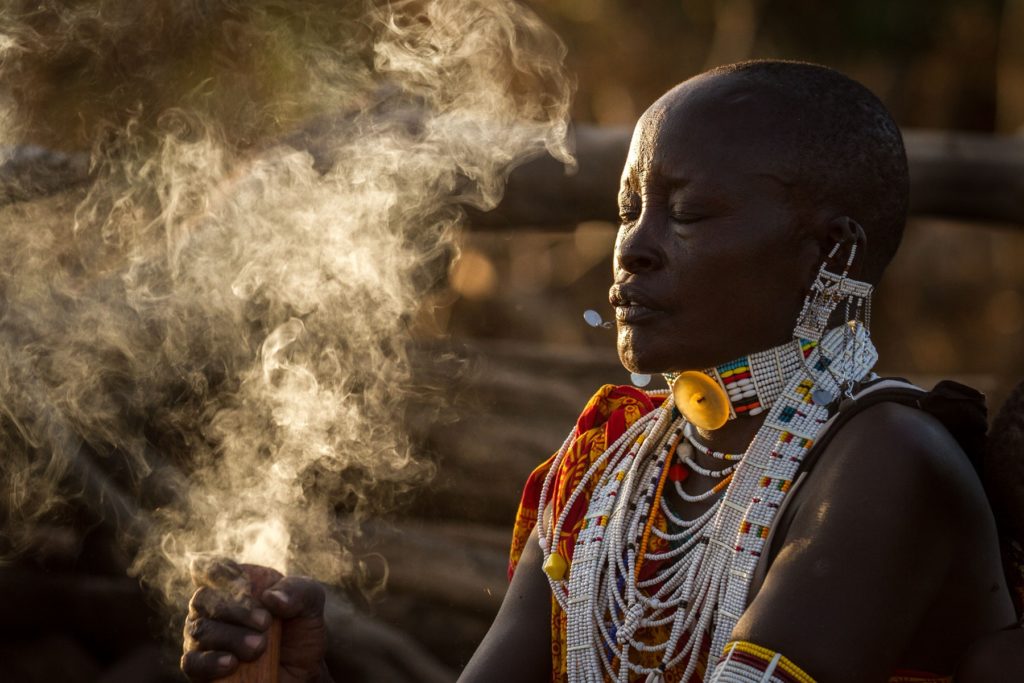 Maasai woman in layered beaded collars and earrings cleans a gourd with smoke, her face lit by low golden light.