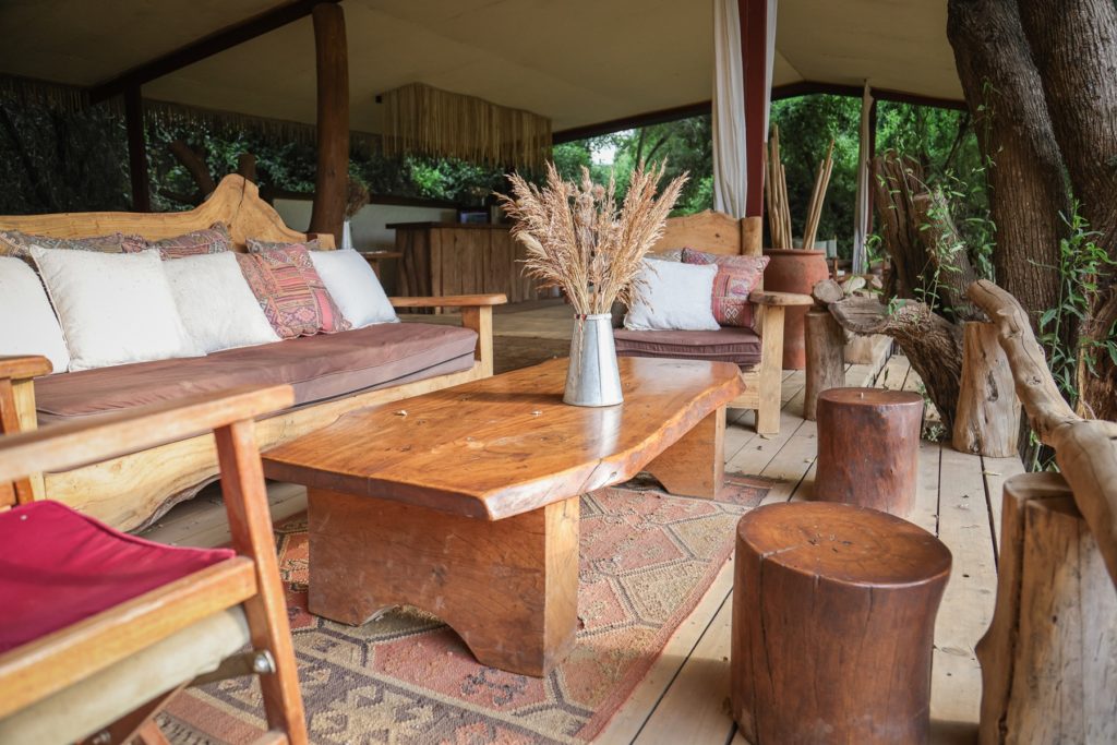 Main lounge at Shompole Wilderness with a live-edge timber table, cushioned wooden sofas, log stools and a kilim rug on the deck.