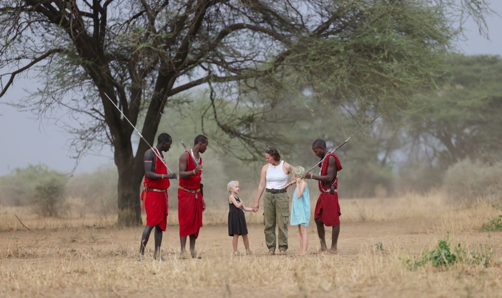 Three Maasai hosts carrying spears talk with a guest and two children under an acacia tree on a hazy day in Shompole Conservancy.