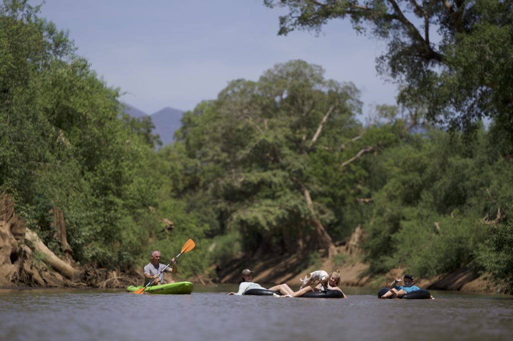 Guests float down a tree-lined river on inner tubes beside a kayaker, with a small dog and a waving child joining in.