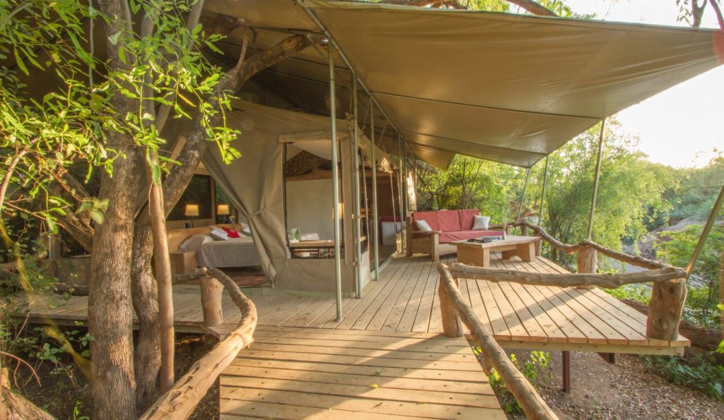 Raised timber deck with branch railings leading to a canvas tent and shaded lounge area among trees in late afternoon light.