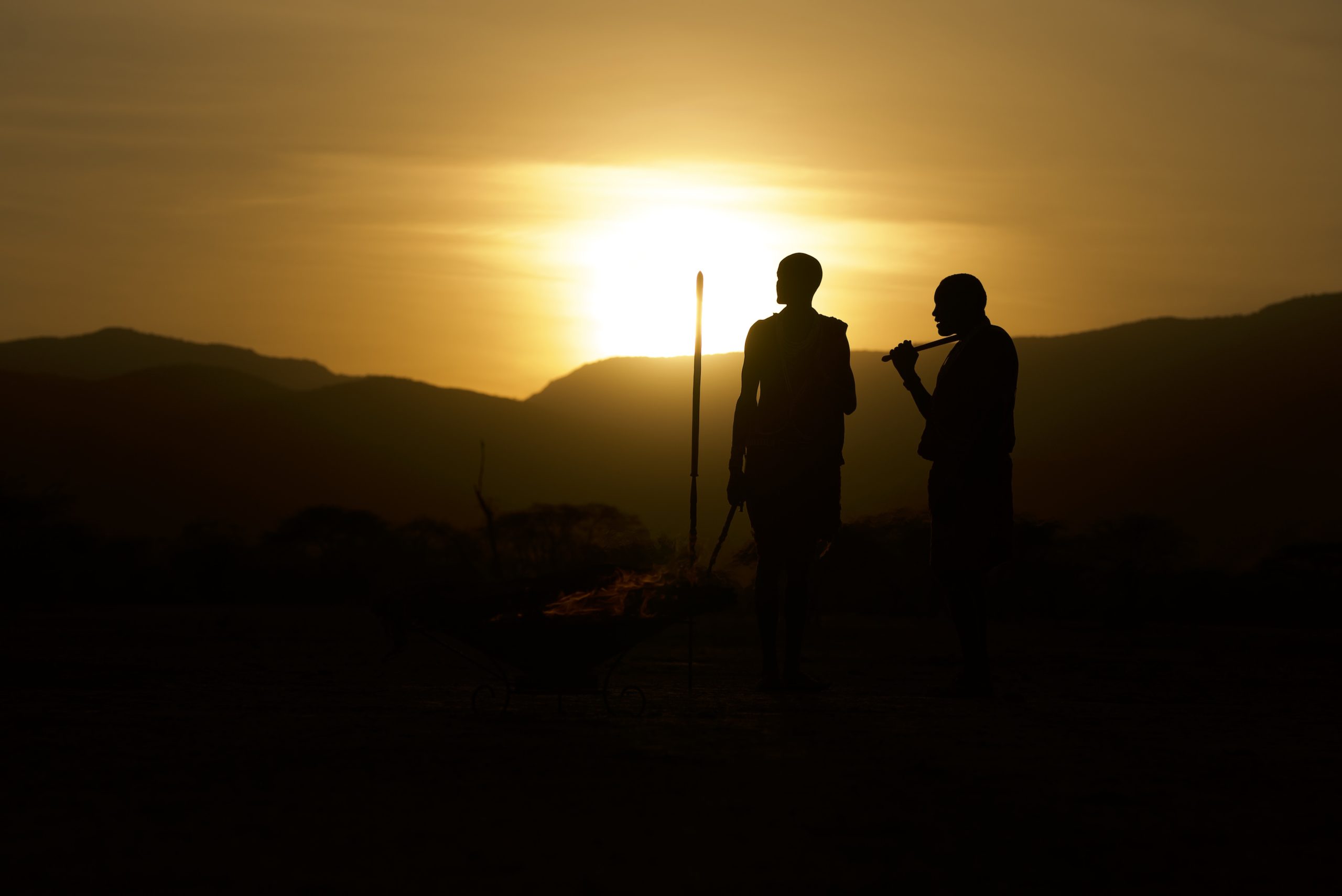 Two Maasai figures silhouetted beside a fire pit at sunset, one holding a spear, with hills fading into golden haze behind.