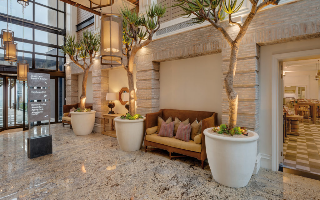 Reception lobby at Strand Hotel Swakopmund with tall aloe trees, brick archways and signage pointing to Ocean Cellar, Brewer & Butcher and the Atlantic Spa