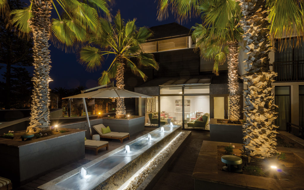 Illuminated palm trees and water feature on a terrace at Strand Hotel Swakopmund by night, with the spa lounge glowing beyond