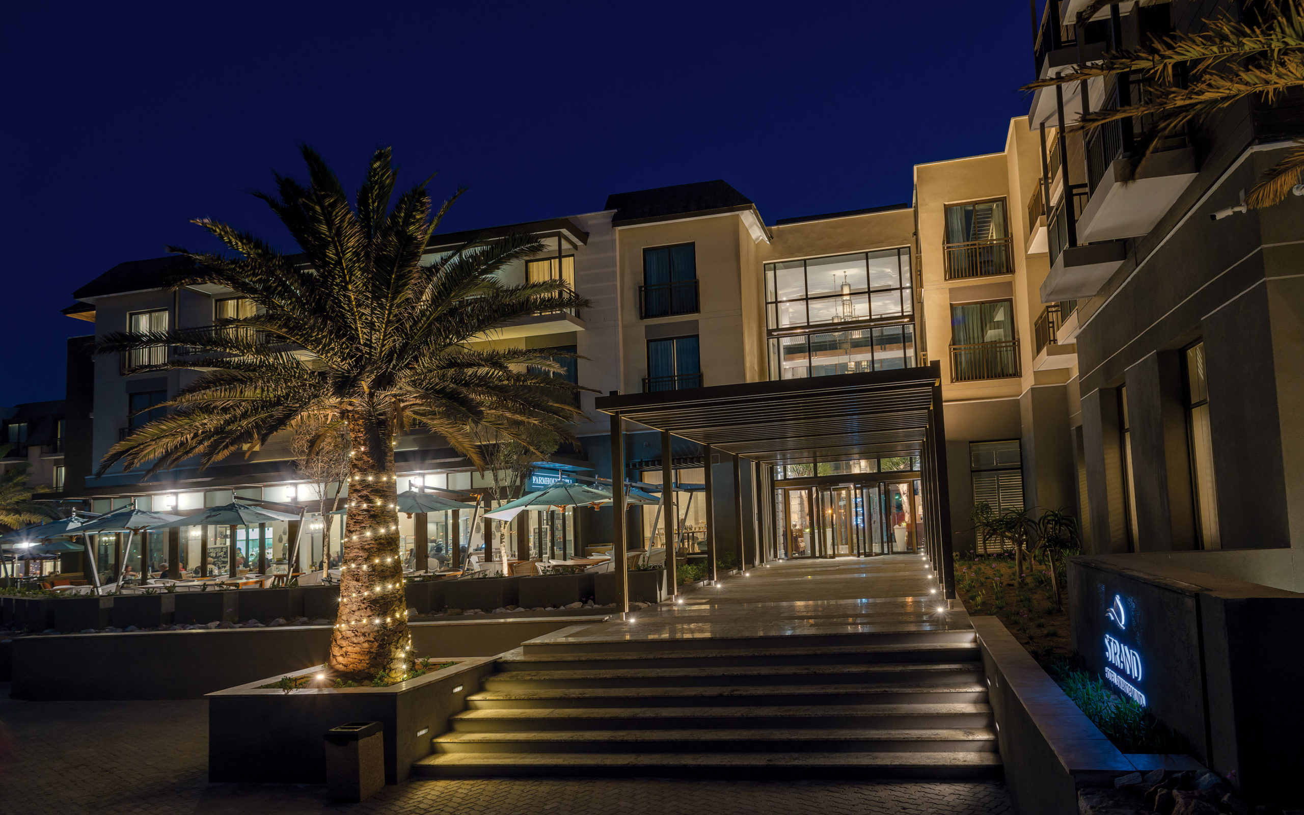 Evening view of the Strand Hotel Swakopmund entrance, lit walkway and palm-lined terrace against the night sky