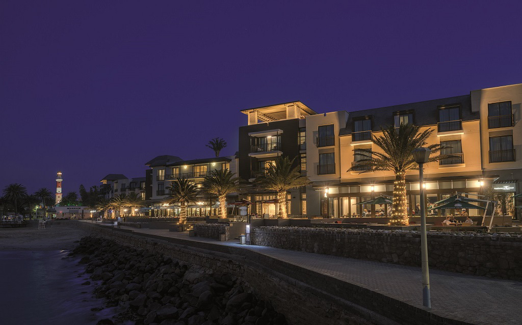 Strand Hotel Swakopmund illuminated at night along the Mole, with the Swakopmund lighthouse visible in the distance