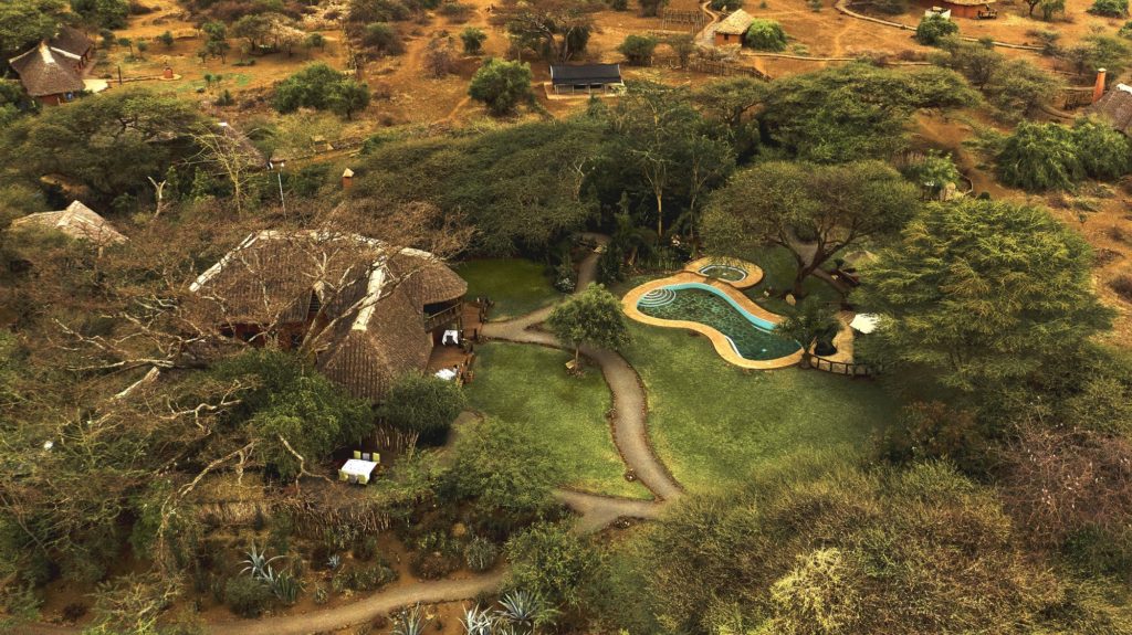 Aerial view of Tawi Lodge's thatched main area, curved swimming pool and lawns among acacia trees, with cottages scattered beyond.