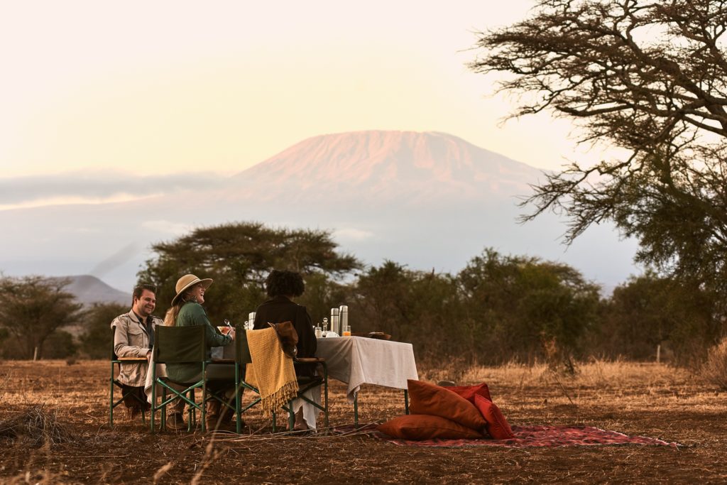 Guests share a bush breakfast at a cloth-covered table on open plains, with Mount Kilimanjaro lit pink at dawn behind.