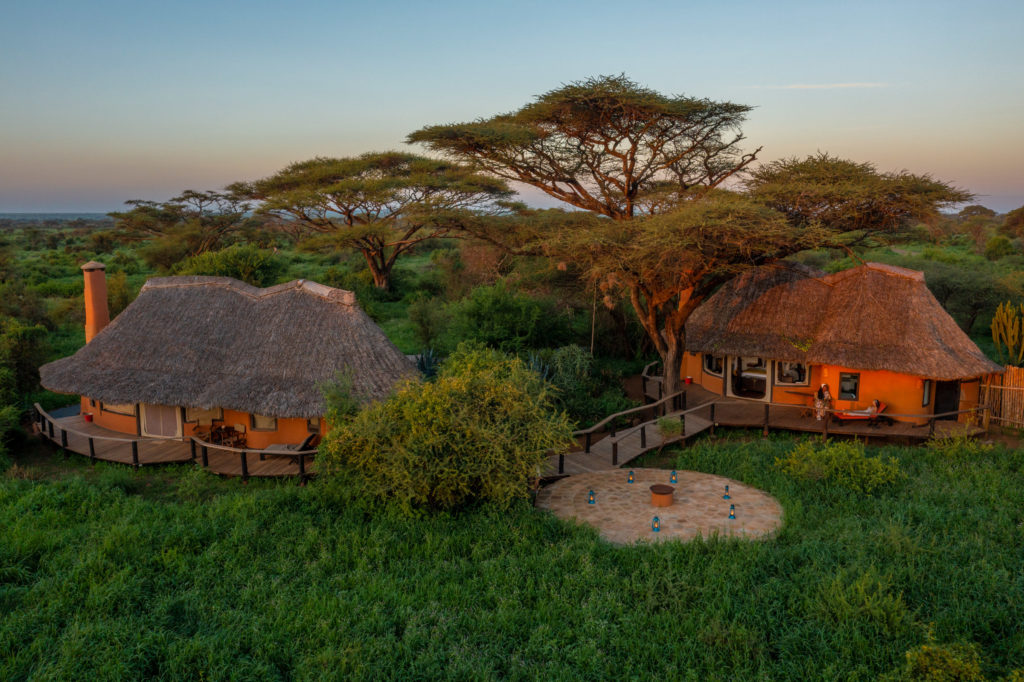 Aerial view of two thatched Tawi Lodge cottages joined by a boardwalk, with a lantern-lit stone fire pit and tall acacias.