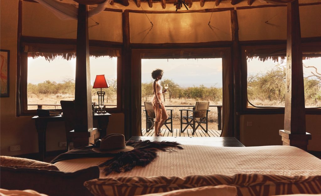 A guest holding a glass of wine walks across the cottage veranda, seen past a four-poster bed and a safari hat inside.