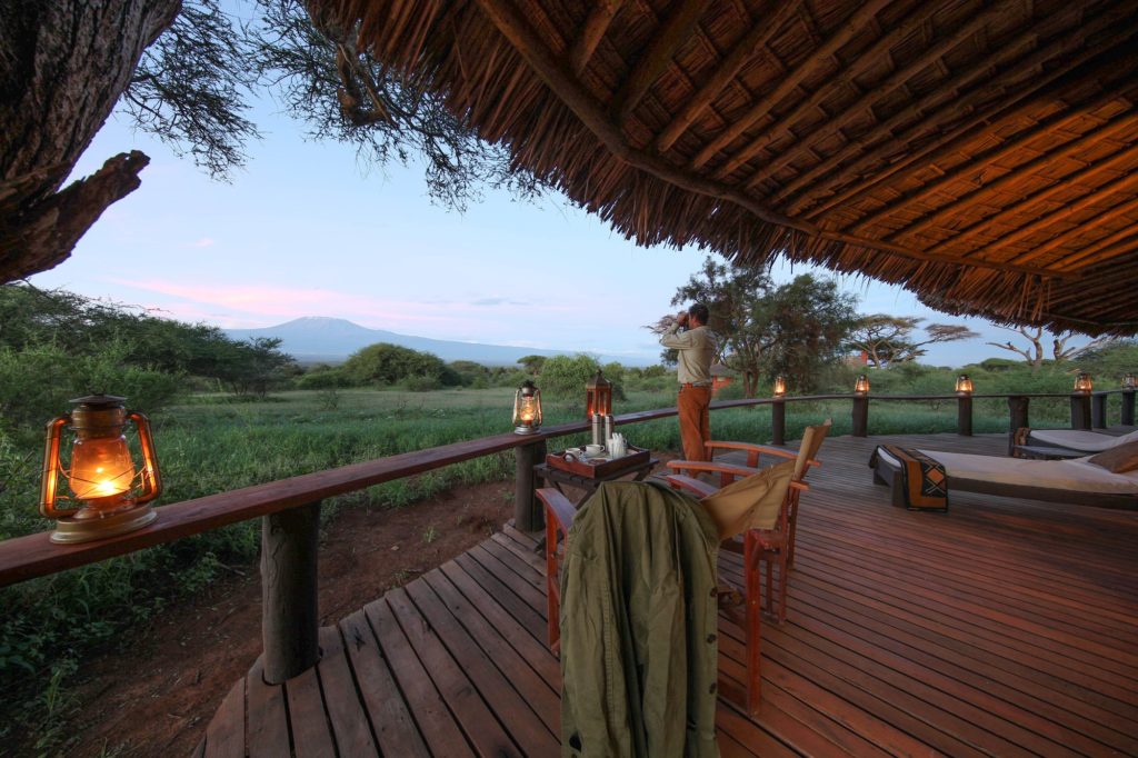 A guest scans the bush with binoculars from a lantern-lit cottage deck at Tawi Lodge, with snow-capped Mount Kilimanjaro beyond.