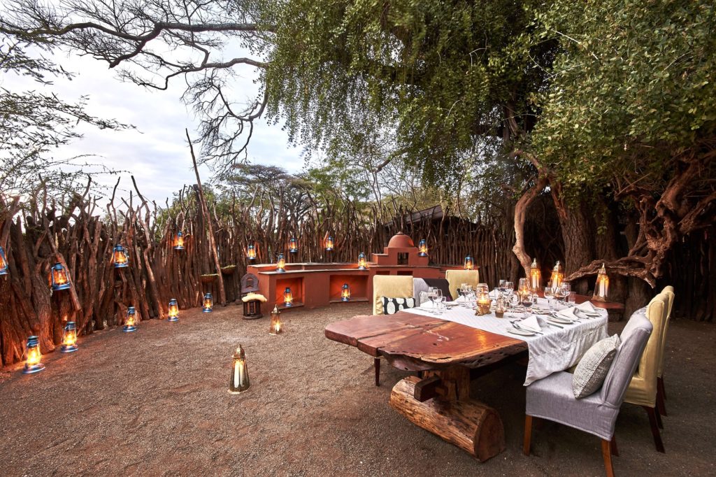 Outdoor dining boma at Tawi Lodge set for dinner, with a live-edge wooden table, lanterns and a stick fence under acacias.
