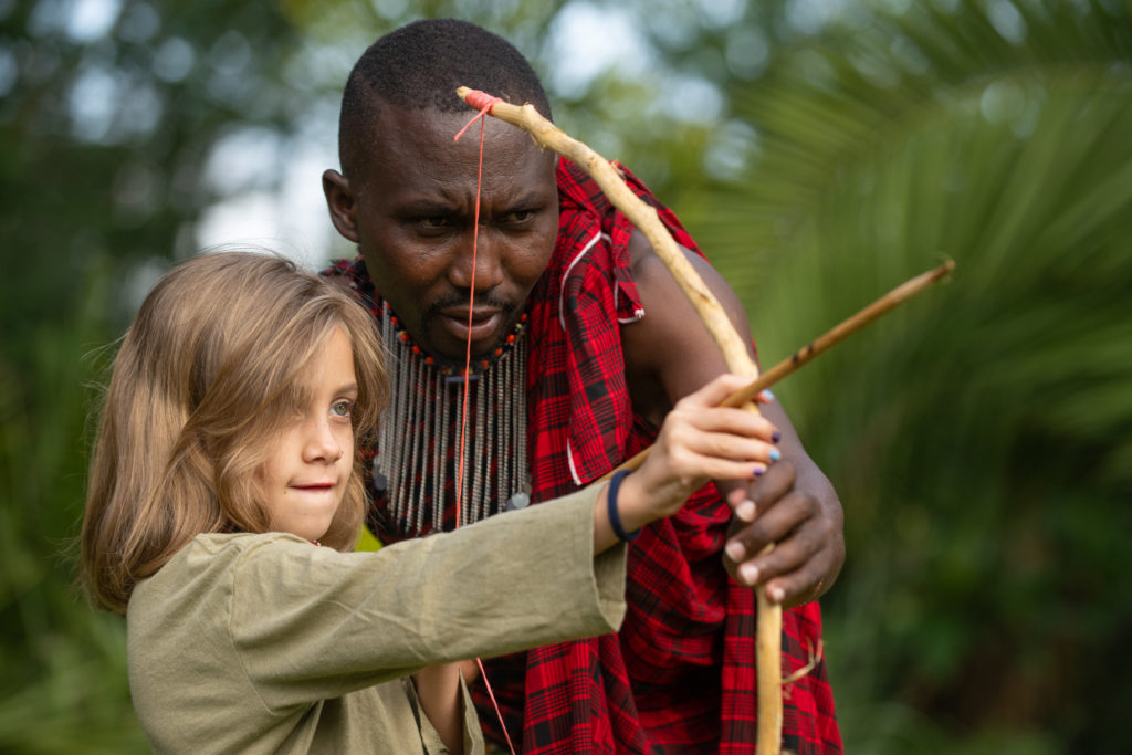 A guide in a red shuka and beaded collar shows a child how to draw a handmade wooden bow and arrow at Tawi Lodge, Kenya.