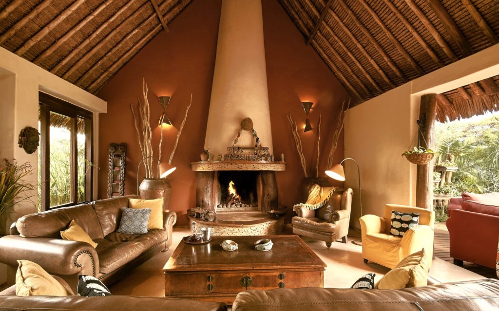 Lounge at Tawi Lodge with a log fire in a stone hearth, leather sofas, a wooden chest coffee table and a high thatched ceiling.