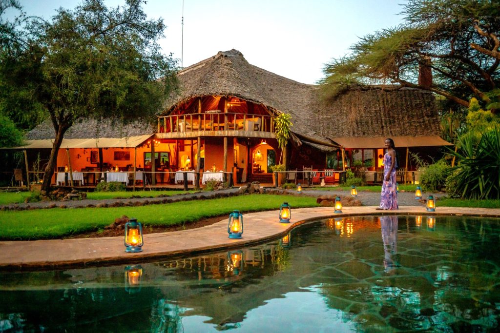 Tawi Lodge's thatched main building lit at dusk, with an upstairs lounge, dining veranda and a lantern-lined pool where a guest stands.