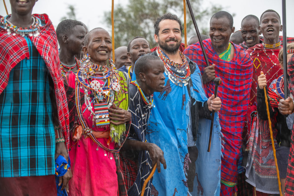 A guest wrapped in a blue shuka joins Maasai community members in beaded jewellery for singing and dancing in light rain.