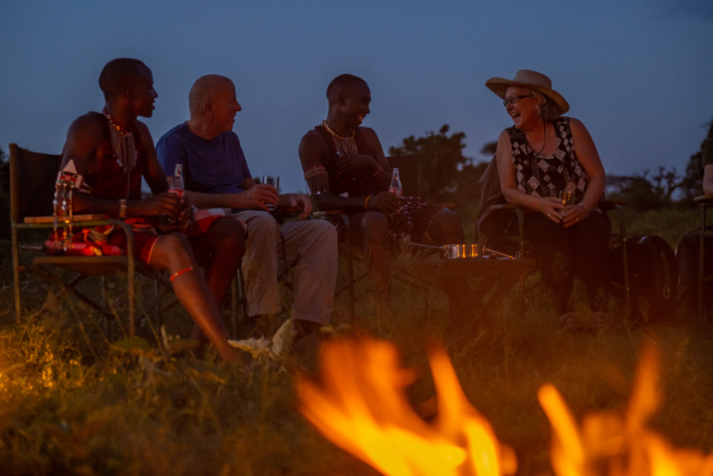 Guests and two guides in traditional beaded dress share drinks around a campfire at sundowner time as the sky turns deep blue.