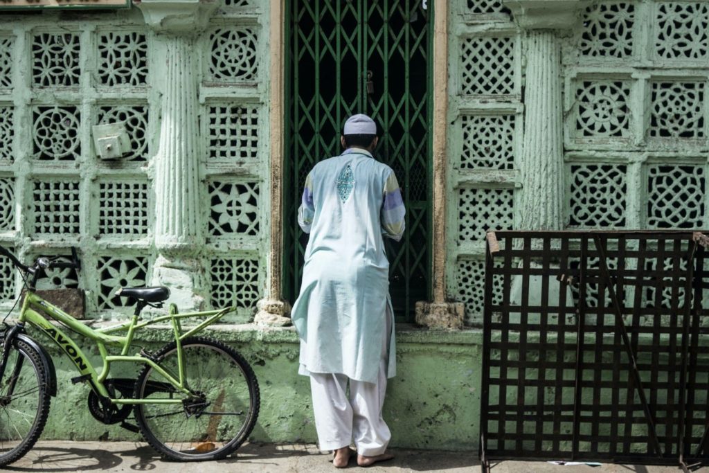 A man in traditional dress stands at a latticed green shutter outside a carved heritage facade in the old city, a bicycle parked nearby.