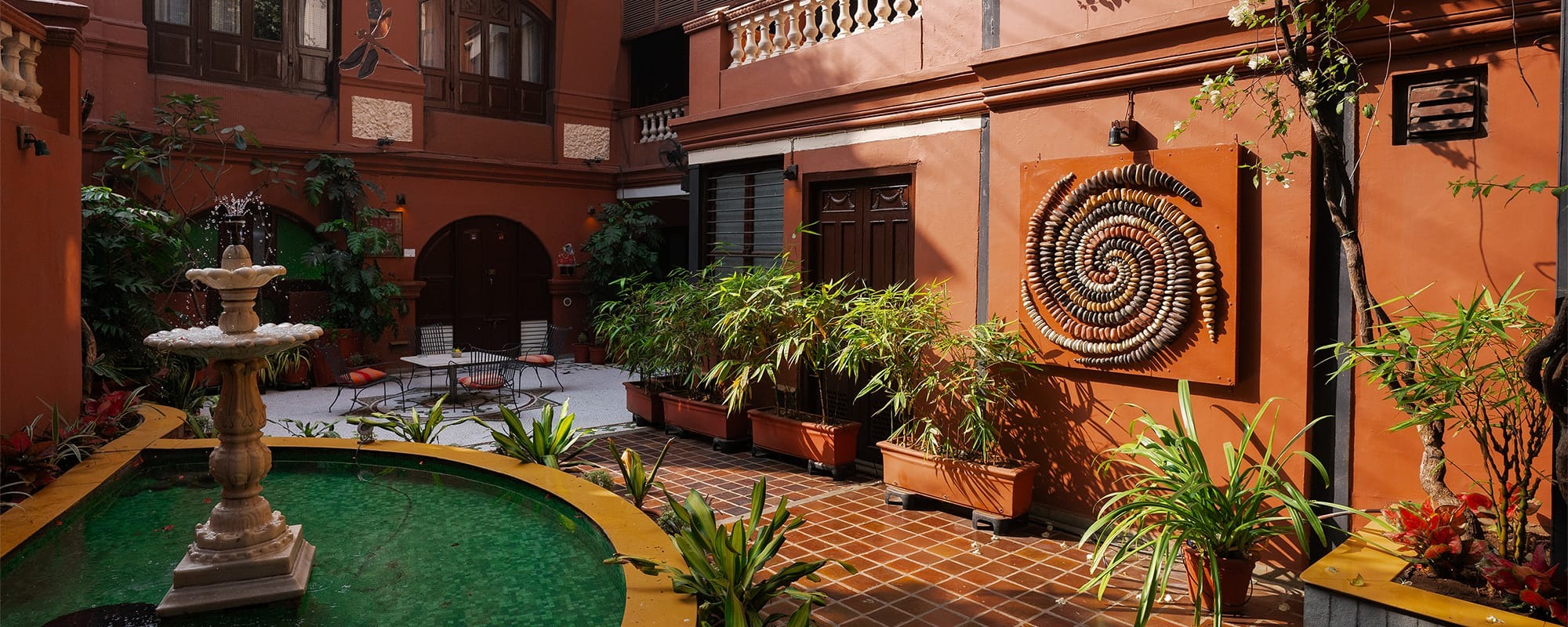 A stone fountain trickles beside a mosaic-tiled courtyard, its terracotta walls decorated with a spiral pebble artwork and potted greenery.