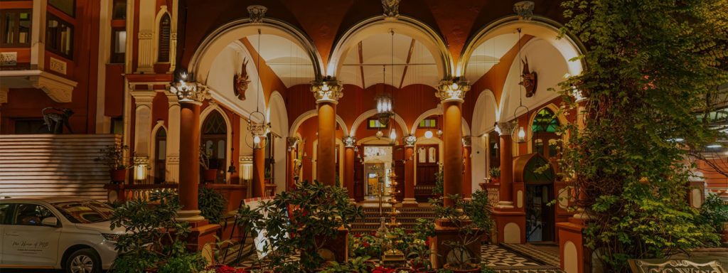 The hotel's arched colonnade entrance glows warmly at night, flanked by carved pillars, hanging lanterns and lush potted plants.