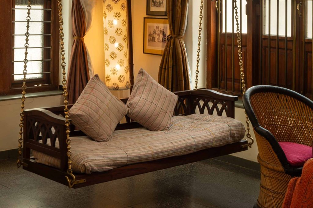 A wooden swing seat draped with striped bolster cushions hangs on brass chains beside a rattan chair and vintage family photographs.
