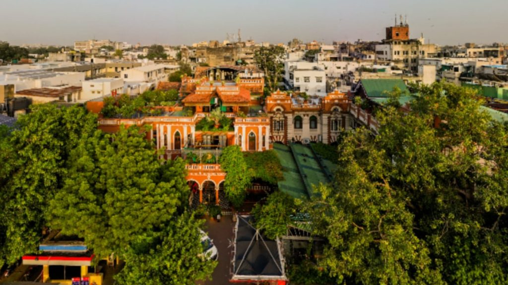 An aerial view at golden hour shows the hotel's terracotta and white facade nestled among treetops within Ahmedabad's old city.