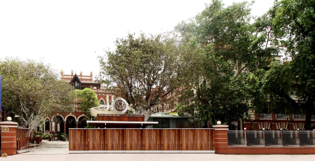 A wooden entrance gate bearing The House of MG's crest sits beneath mature trees, with the hotel's arched red facade visible beyond.