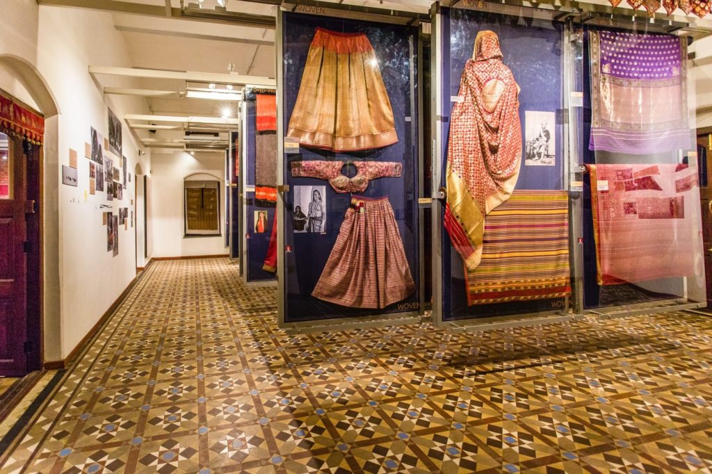 Antique embroidered garments and textiles hang under glass in the heritage gallery, lit against deep blue display panels.
