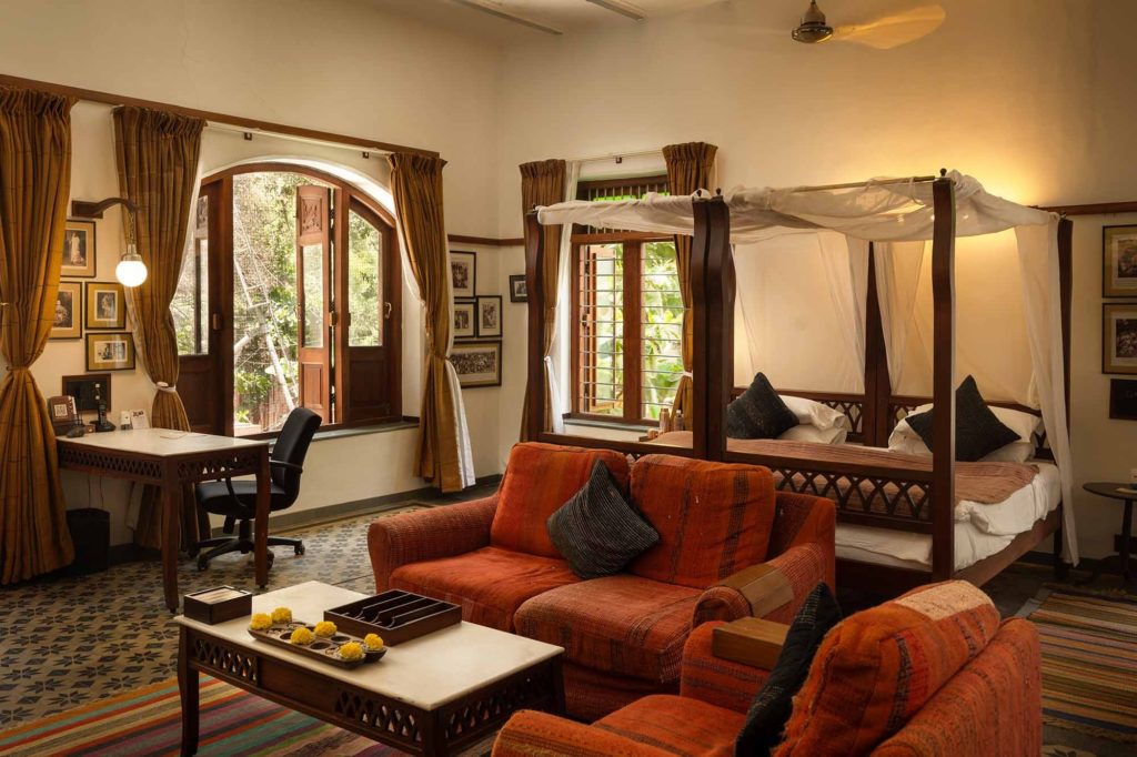 A four-poster bed draped in white canopy fabric sits beside a rust-orange sofa and writing desk in one of the hotel's heritage-styled suites.