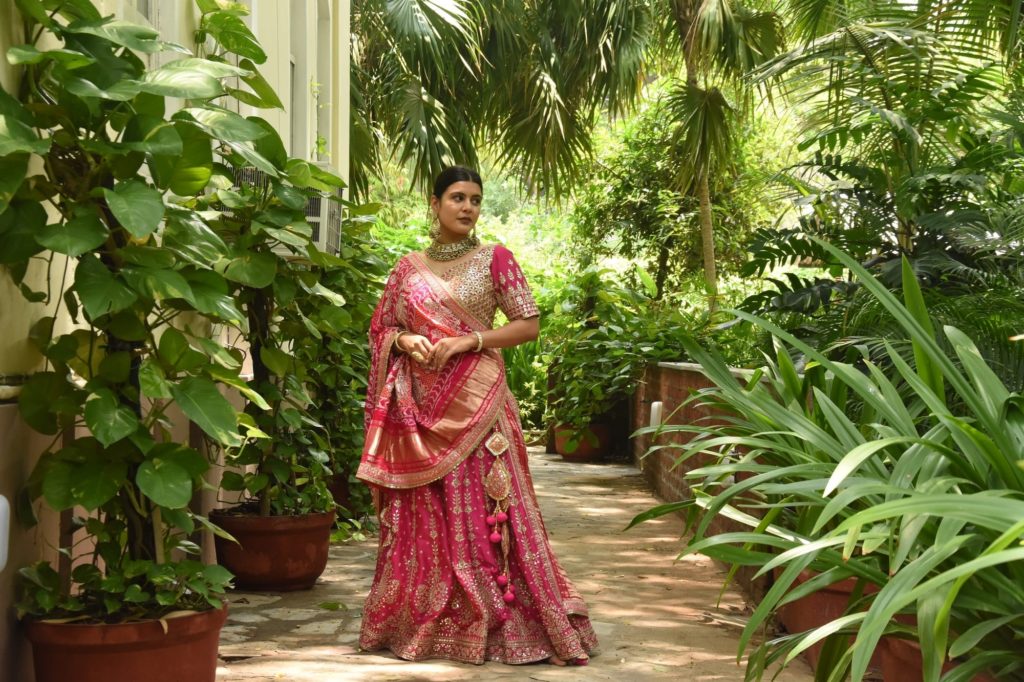 A woman in a pink and gold embroidered sari walks along a garden path shaded by palm fronds and lush potted greenery.