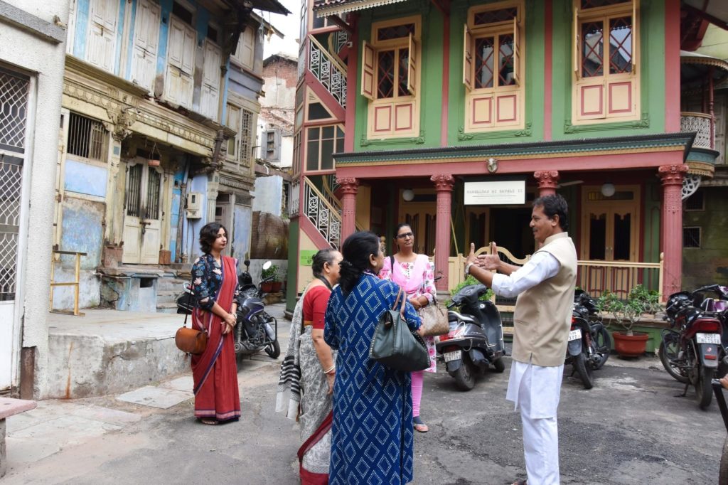 A guide gestures while addressing a small group of guests on a heritage walk outside the colourfully painted Mangaldas ni Haveli.