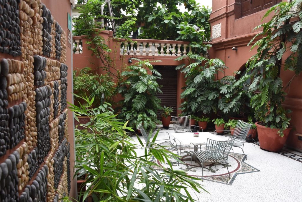 A pebble-tiled wall borders a leafy courtyard furnished with metal chairs and a mosaic-topped table.