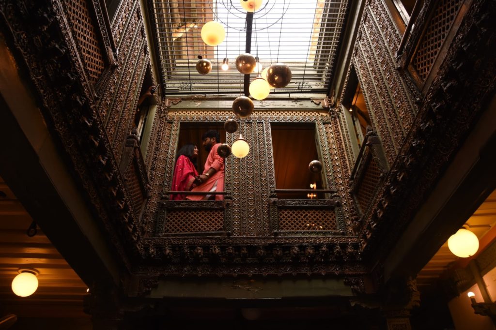 A couple stands on a carved wooden balcony beneath hanging globe lights within the hotel's ornate central atrium.
