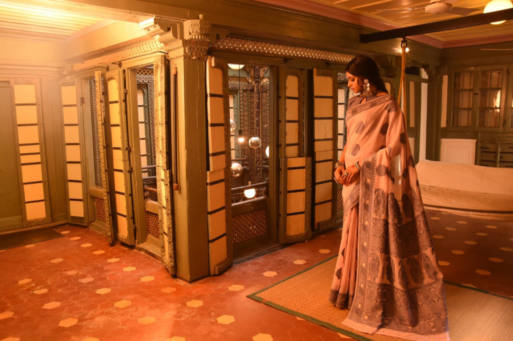 A woman in a dusky pink printed sari stands beside an antique wooden almirah in a warmly lit heritage interior.