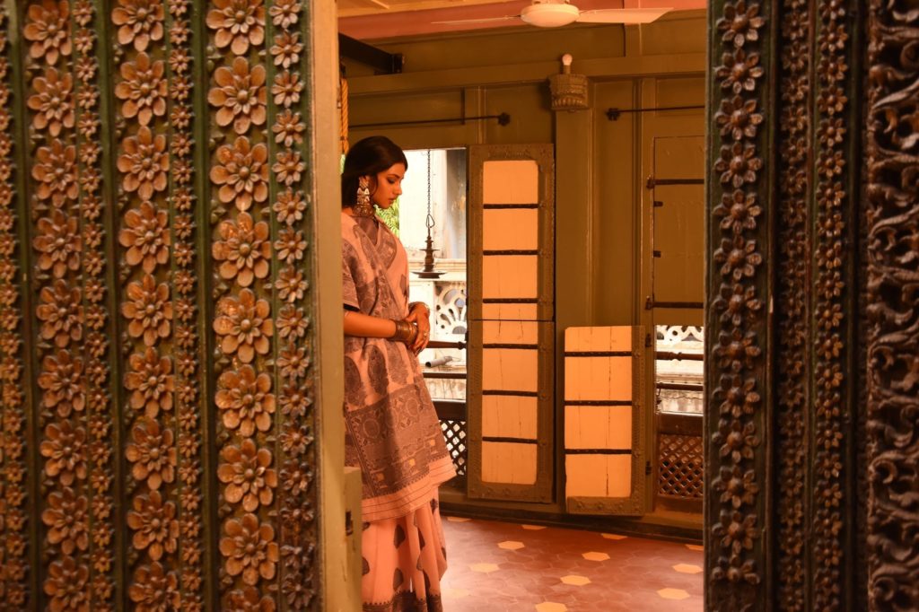 A woman in a printed sari stands framed within an intricately carved wooden doorway in a warmly lit heritage interior.