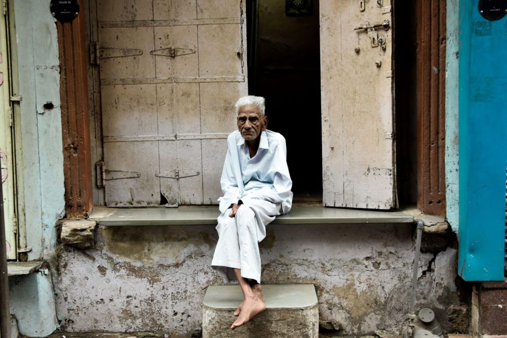 An elderly man in white sits barefoot on a stone doorstep between the weathered wooden doors of an old city house.