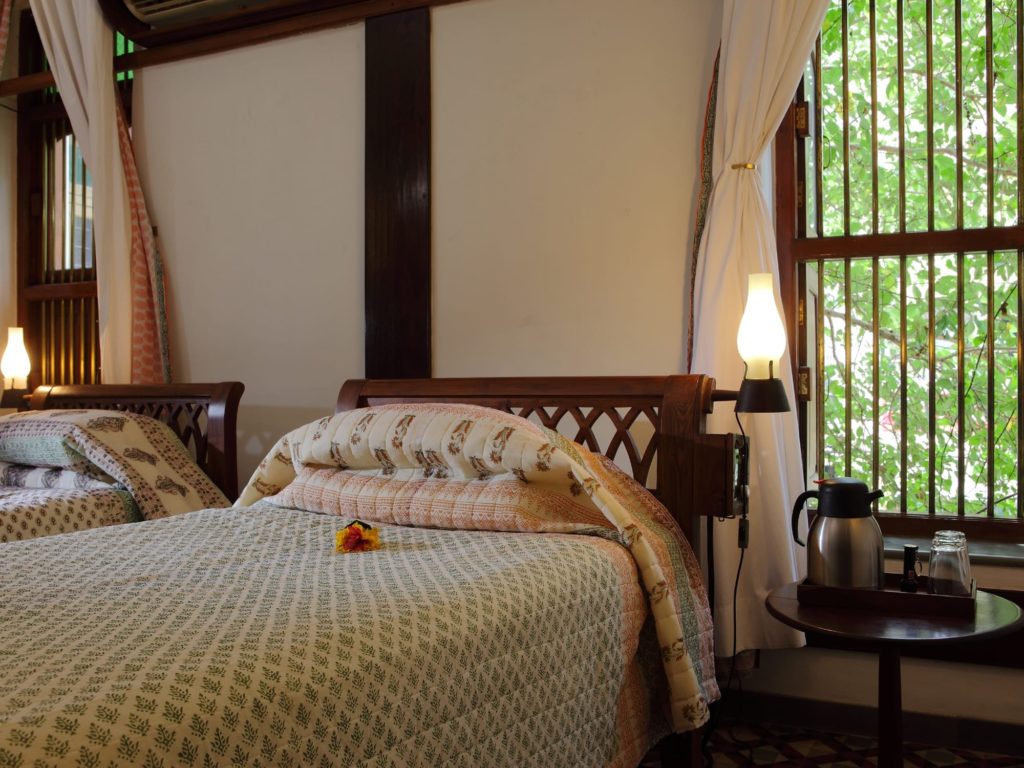 Twin beds dressed in block-printed quilts sit beneath tall barred windows overlooking green foliage, with a brass coffee set on a bedside table.