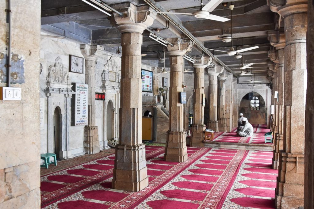 Sunlight filters through the marble colonnade of the Jama Masjid, where two men sit in quiet prayer on red patterned carpets.