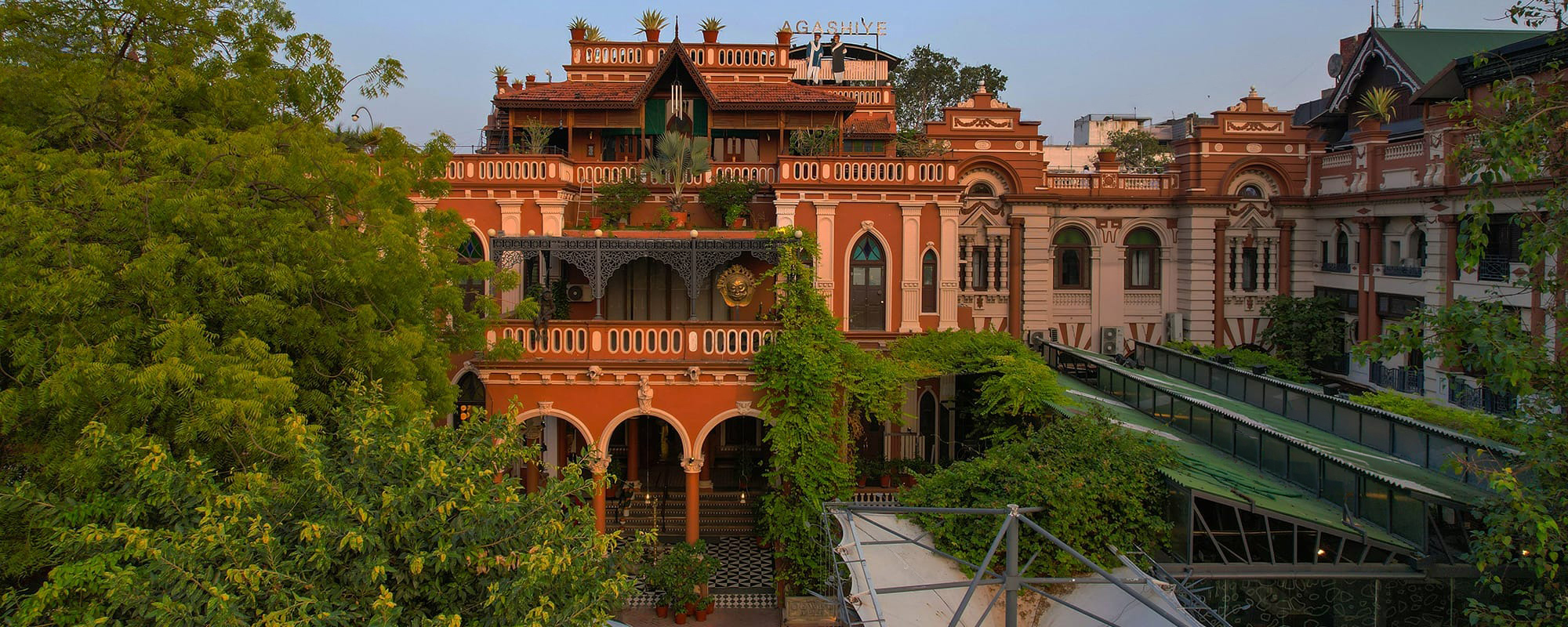 At golden hour, the hotel's terracotta and cream facade rises above lush greenery, the rooftop Agashiye restaurant and Green House café signage visible below.