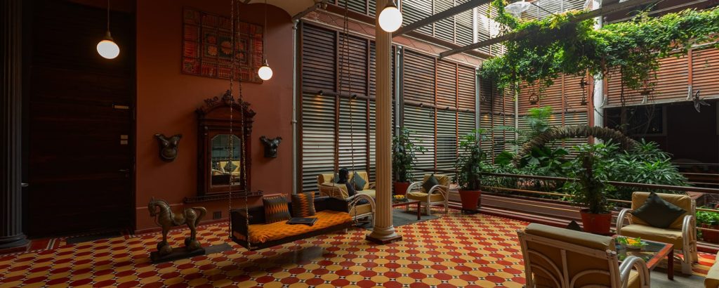 A skylit lounge with checkerboard tile flooring features a hanging swing seat, rattan armchairs and trailing greenery along a glass-panelled wall.