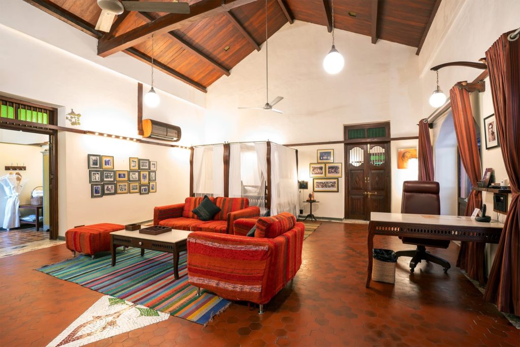 A spacious suite features rust-red sofas, a sheer-curtained four-poster bed and a wall of black-and-white family photographs beneath a peaked wooden ceiling.