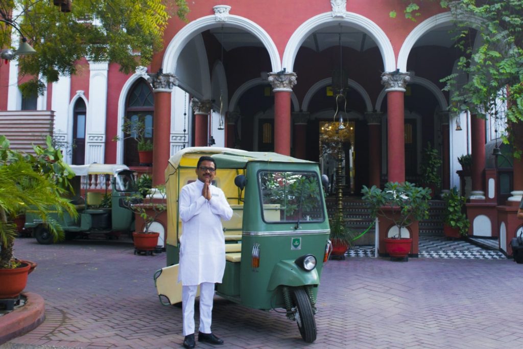 A hotel host in white kurta stands beside a vintage green rickshaw beneath the red and white colonnaded facade of The House of MG's entrance.